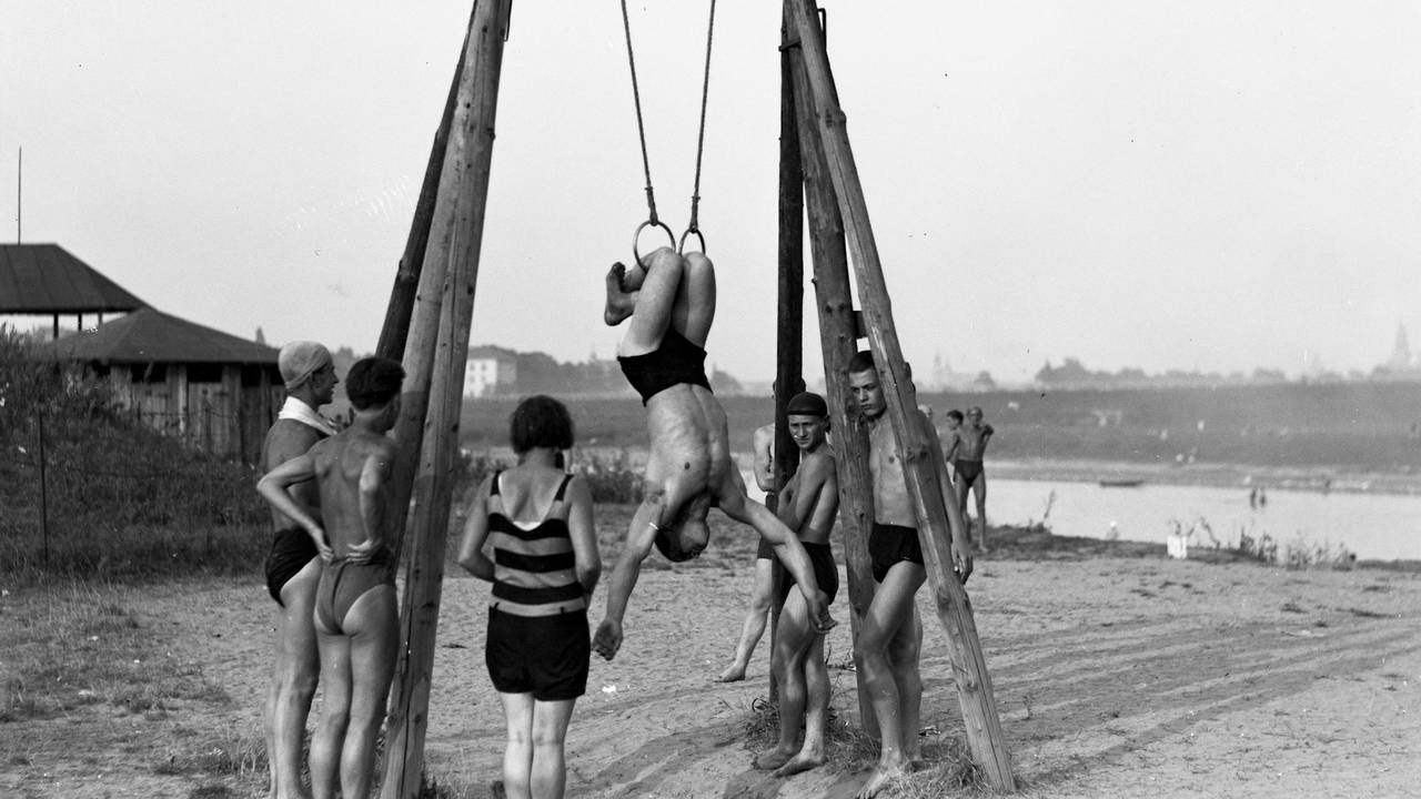 Fotografia pozioma, czarno-biała. Kompozycja otwarta. Krajobraz plaży przy rzece. Grupa ludzi w kostiumach kompielowych w stylu retro obserwuje ćwiczenia mężczyzny na kółkach gimnastycznych. 